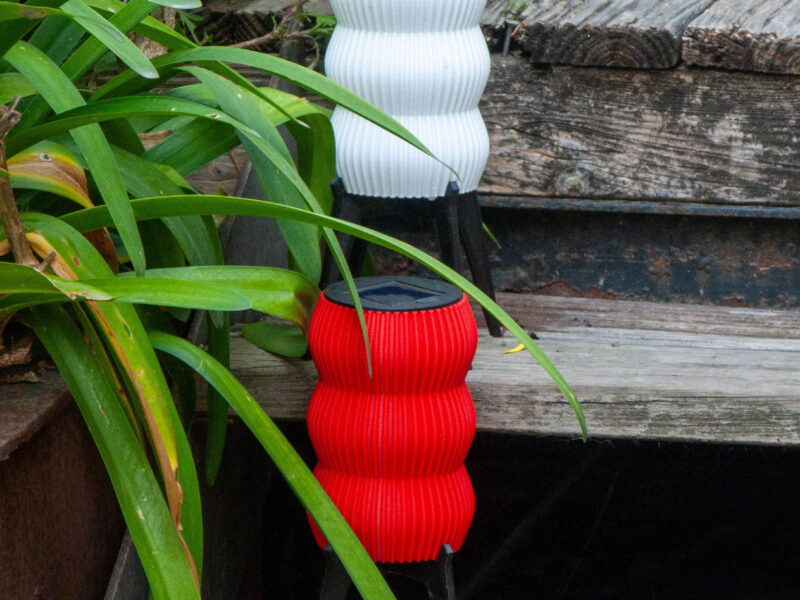 Deux lampes solaires de jardin "GUINGET" imprimées en 3D, une blanche et une rouge, en plastique recyclable, posées sur des marches en bois entourées de plantes.