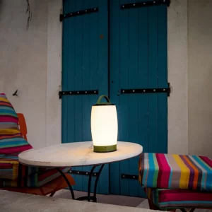 Une lampe LED allumée sur une table ronde, entourée de deux chaises avec des coussins colorés devant des volets bleus.