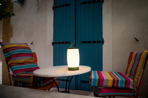 Une lampe LED allumée sur une table ronde, entourée de deux chaises avec des coussins colorés devant des volets bleus.