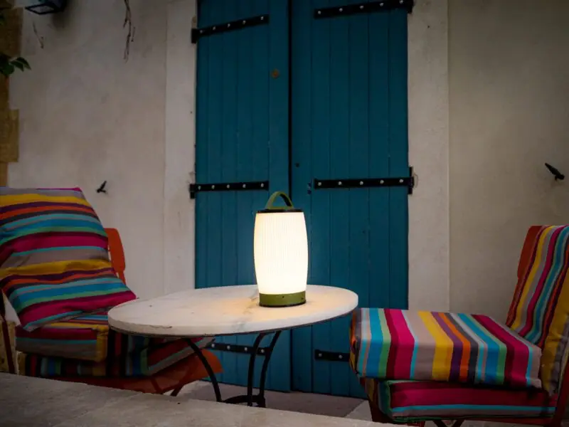 Une lampe LED allumée sur une table ronde, entourée de deux chaises avec des coussins colorés devant des volets bleus.