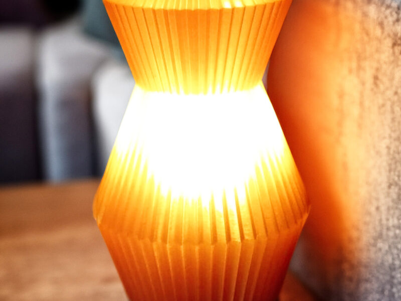 Lampe Voyager orange imprimée en 3D avec un design plissé, posée sur une table en bois près d'un canapé.