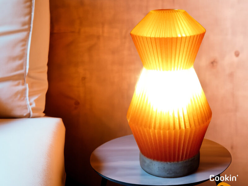 Lampe Voyager orange imprimée en 3D avec un design plissé, posée sur une table d'appoint près d'un lit.