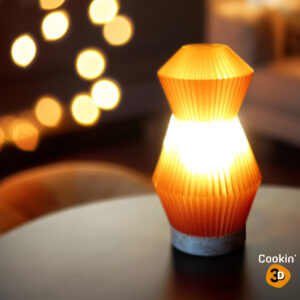 Lampe Voyager orange imprimée en 3D avec un design plissé, éclairant une table dans une ambiance chaleureuse avec des lumières floues en arrière-plan.