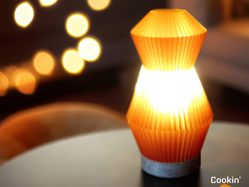 Lampe Voyager orange imprimée en 3D avec un design plissé, éclairant une table dans une ambiance chaleureuse avec des lumières floues en arrière-plan.