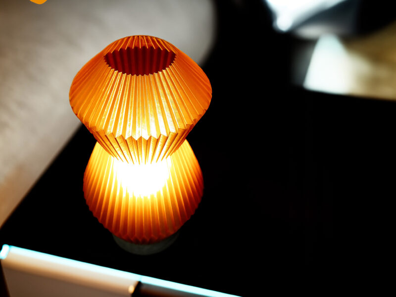 Vue de dessus de la lampe Voyager orange imprimée en 3D avec un design plissé, éclairant une table noire dans une pièce sombre.