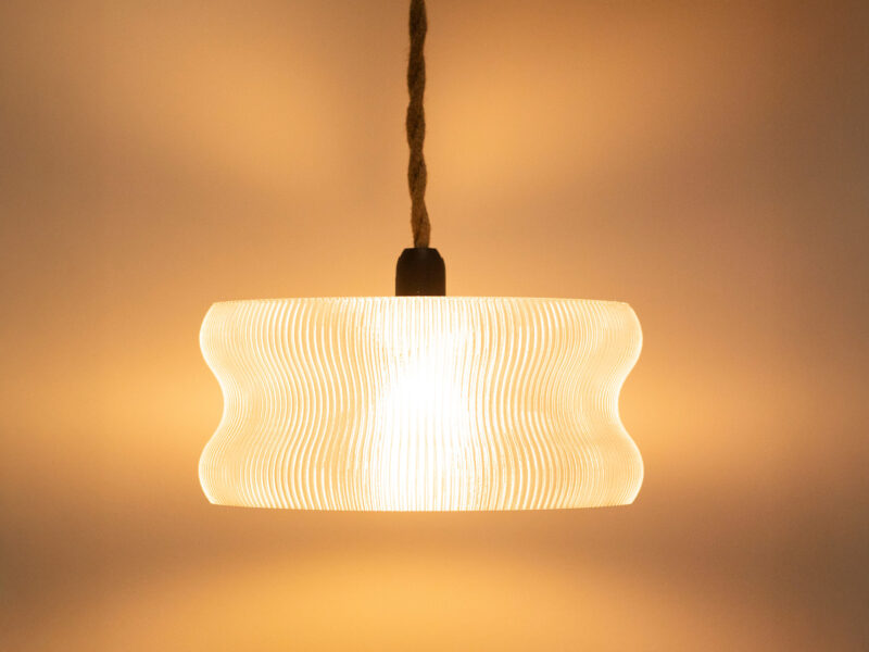 Suspension design en impression 3D avec un abat-jour transparent en forme de cloche ondulée diffusant une lumière chaude.